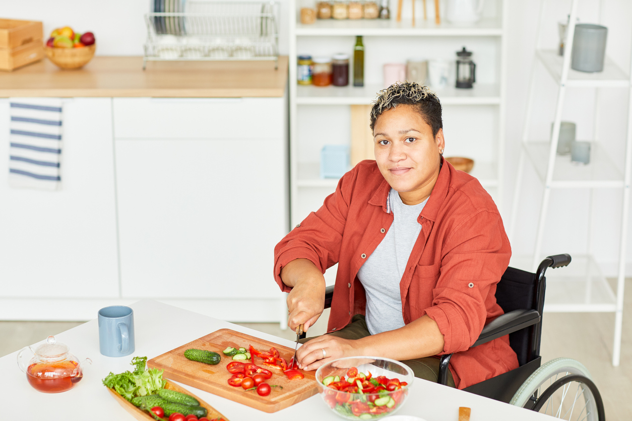 Portrait of African disabled young woman with short hair looking at camera while sitting in wheelchair and cutting vegetables in the kitchen