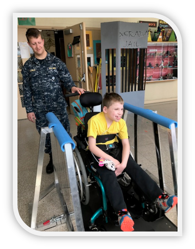 Navy sailor wearing blue camouflage behind Brennon in his wheelchair on glider.