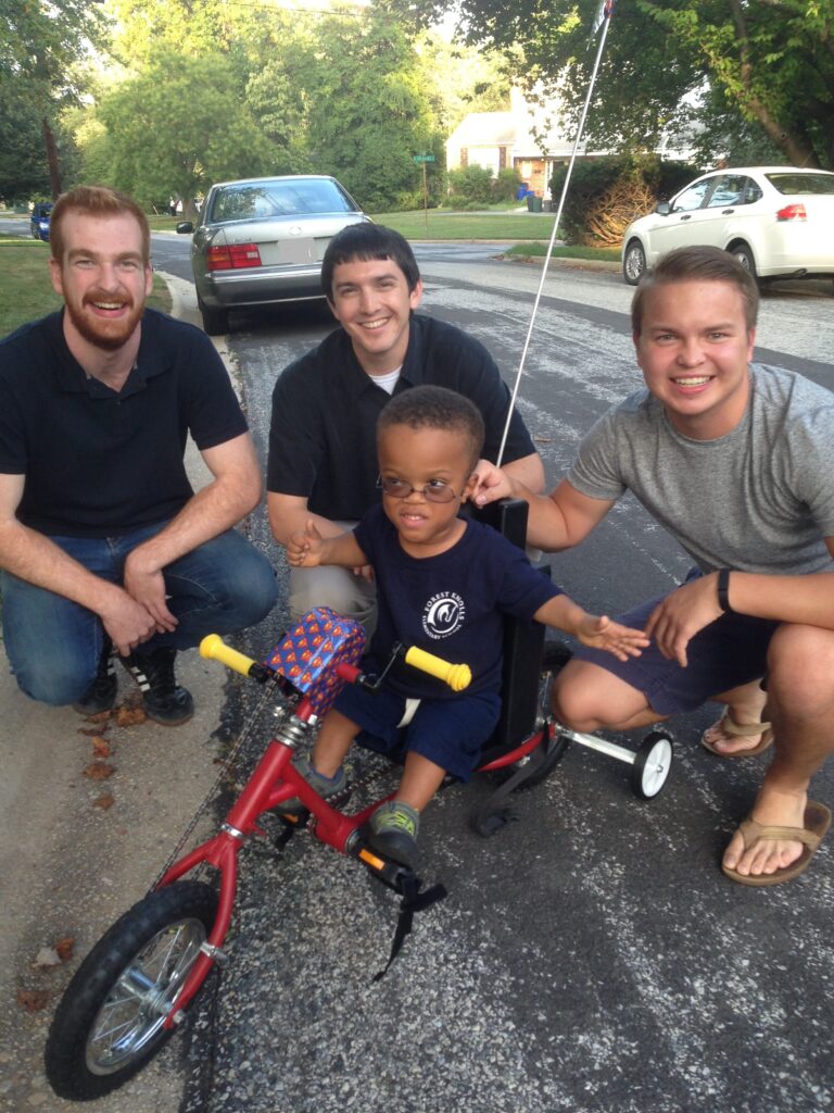Three smiling engineers with a young boy excited to use his new adapted bicycle.