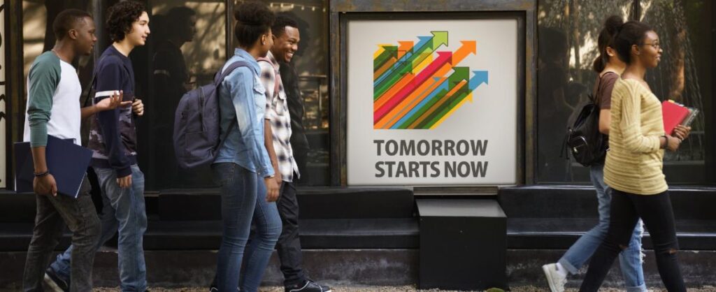 Students walking to the right down a sidewalk with books. They are walking in front of a sign with arrows pointing upwards that reads "Tomorrow starts now". 