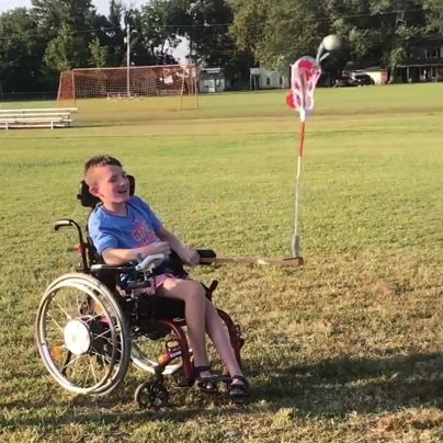 Boy in a motor wheelchair with an engineered lacrosse stick attached to the left arm featuring a lever to catapult the ball.