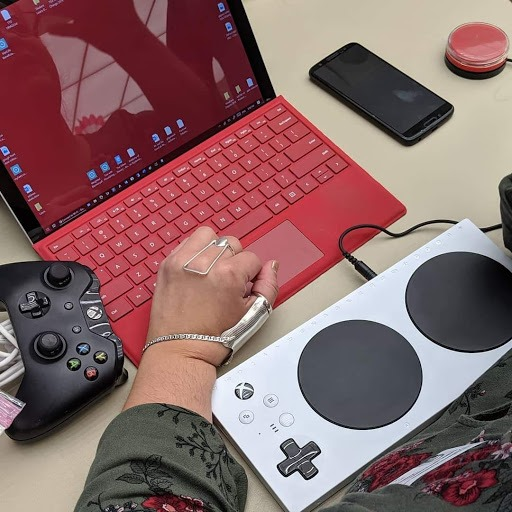 Person's hand wearing adaptive equipment with a red gamers laptop connected to an adaptive x-box panel controller along with a cell phone andstandard x-box controller.