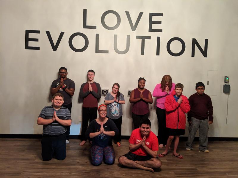 Students pose together in a Namaste pose in front of a wall that says "Love Evolution". 