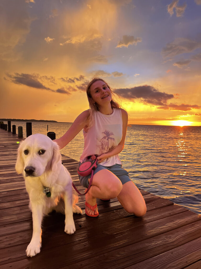 Female student Lily Felton knealing on a dock on the water. The sun is setting behind her as she smiles and holds on to her dog. A golden retriever. 
