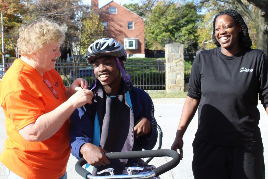 A VME Volunteer helps a child with a disability put on a bicycle helmet as he sits on his new customized adaptive bicycle. His mother smiles behind them.