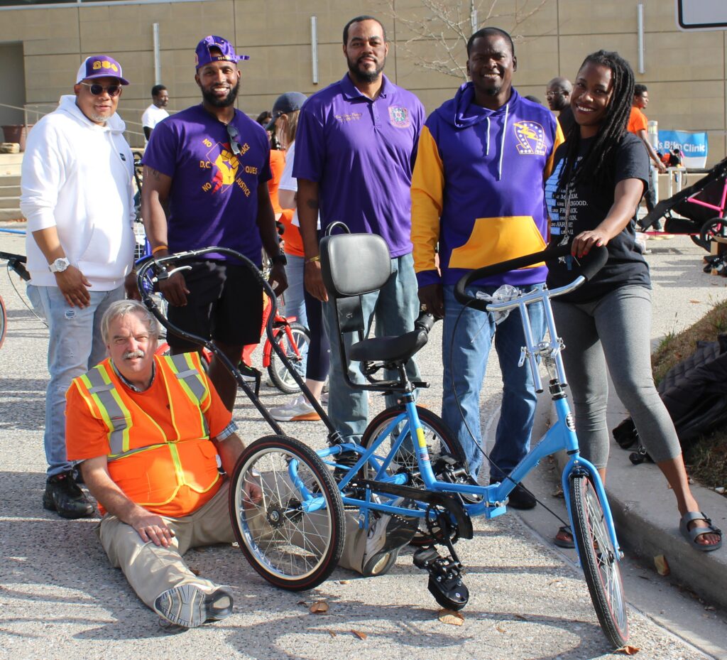 A group of five volunteers smiling around a custom designed adaptive bicycle.