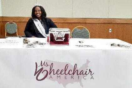 Chandra Smith wearing a tiara and sash at a table that says, "Ms. Wheelchair America."