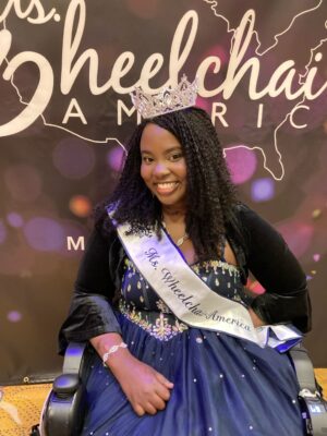 Chandra Smith is a young Black woman with braids. She smiles in front of a sign that says, "Ms. Wheelchair America," and is wearing a glittering tiara and sash.