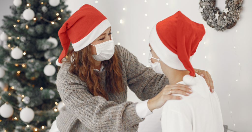 Two women wearing santa hats and face masks. A Christmas tree and wreath are in the background.