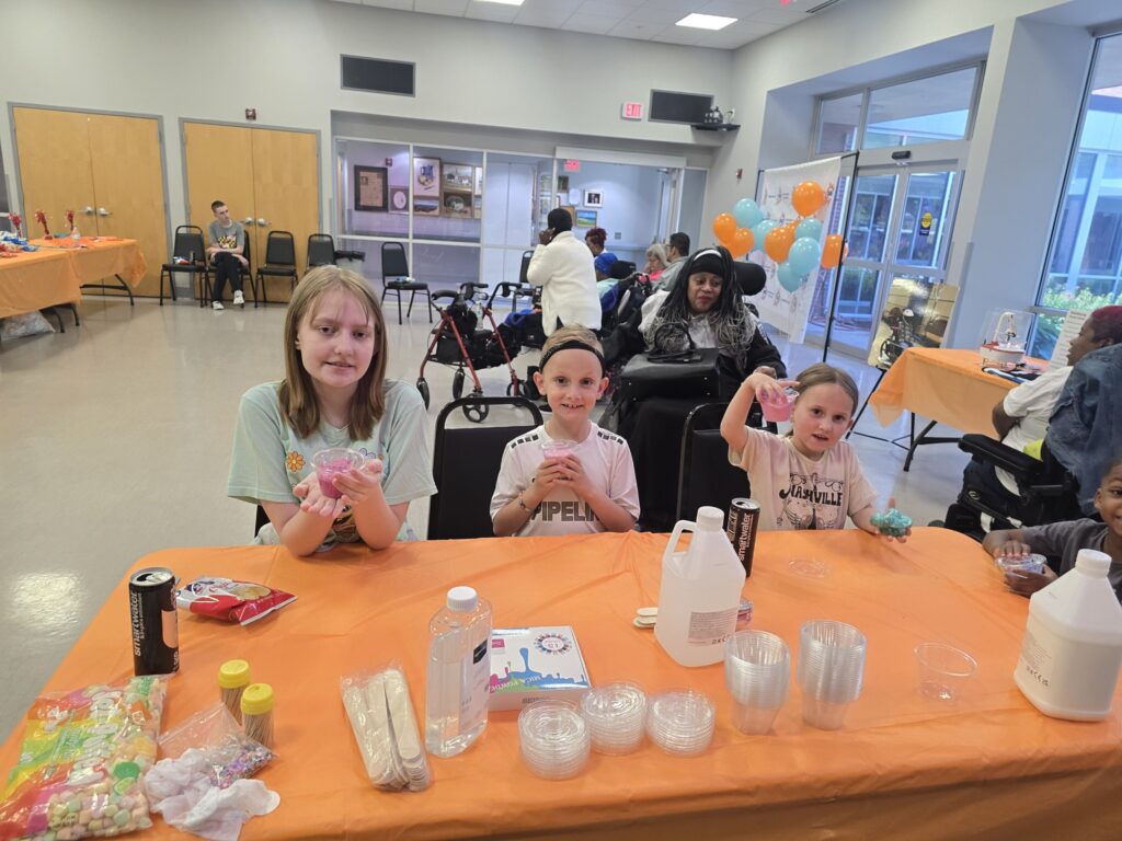 "Three children at the slime table smile and hold up their creations with balloons and other guests in the background."