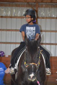 Close-up of a young rider sitting confidently on a black horse, wearing a helmet and glasses. The rider is supported by an adult just outside the frame. The background shows wooden paneling and a purple exercise ball.