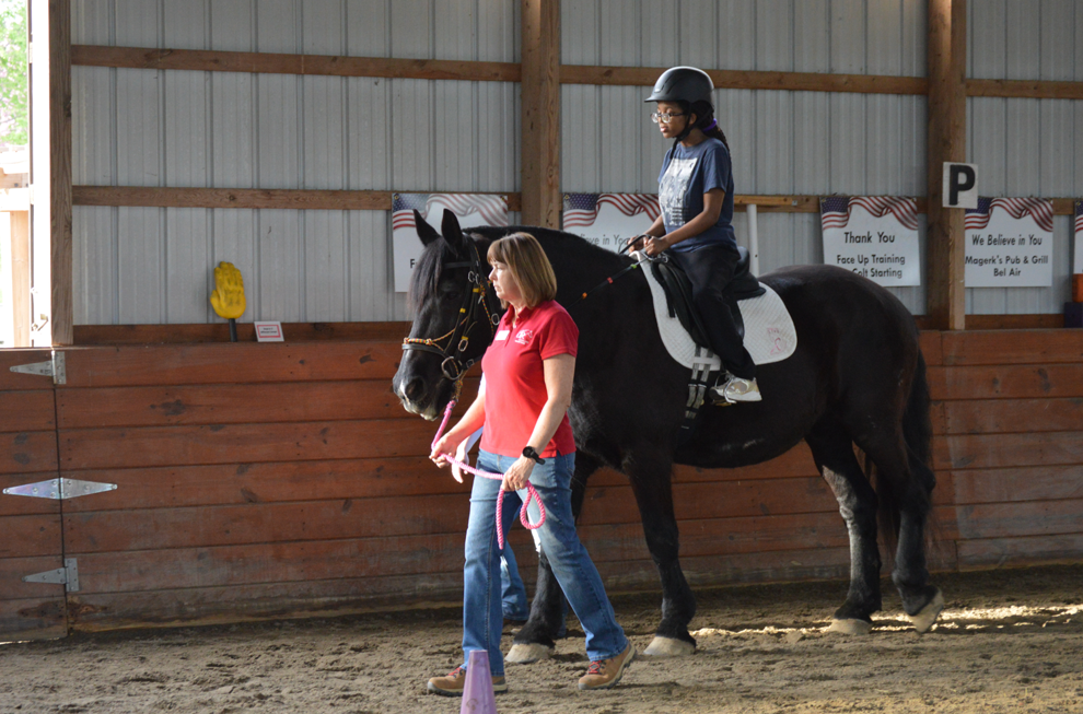A young rider wearing a helmet is guided on horseback by an instructor in a red shirt. The horse and rider are inside a covered arena with wooden walls and American flag-themed signage in the background.