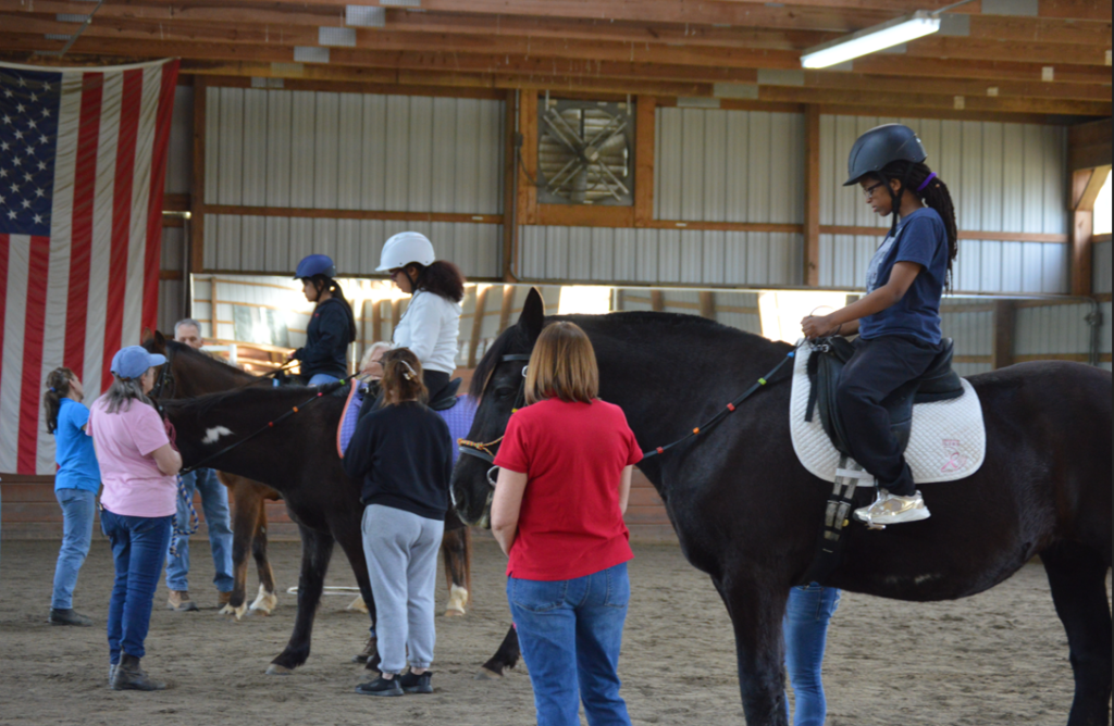 Several riders on horseback participate in a therapeutic riding session indoors. Instructors and volunteers assist as they guide the horses. An American flag hangs on the wall behind them.