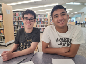 Cole Whitehurst and Glenn Ison smile at a table in the library with pens and worksheets in front of them. Shelves of colorful books are visible in the background.