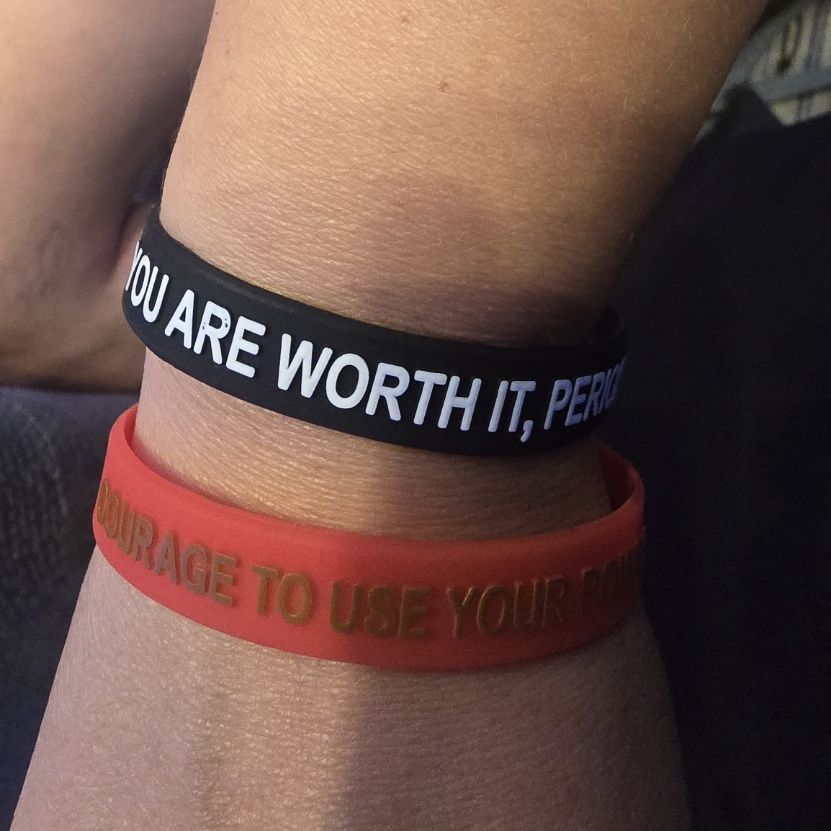 Two silicone wristbands on a person’s wrist, one black with white text reading “YOU ARE WORTH IT, PERIOD” and one red with gold text reading “COURAGE TO USE YOUR POWER.”