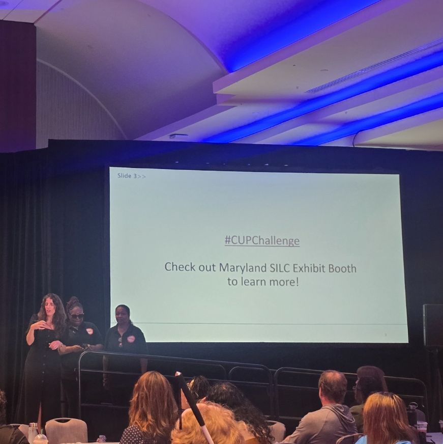 A conference presenter stands on stage beside a large screen displaying the words “#CUPChallenge Check out Maryland SILC Exhibit Booth to learn more!” as attendees watch.