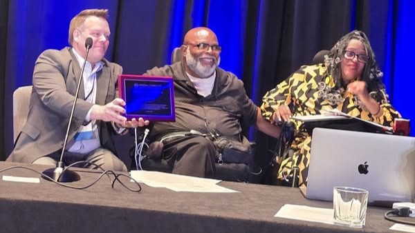 At the front of a stage Janice Jackson is presented a plaque while sitting beside Theo Braddy and another presenter